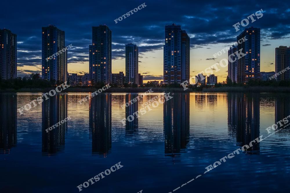 Россия, Санкт-Петербург . Атмосферная фотография вечернего города с домами, расположенными на берегу пруда, где отражения зданий в воде создают эффект зеркального спокойствия. Ночная подсветка и мягкое освещение улиц подчёркивают красоту городского пейзажа, архитектурные линии домов и атмосферу вечернего мегаполиса. Фотография идеально подходит для публикаций о городской жизни, архитектуре, ночных городских пейзажах, туризме и вечерних прогулках по городским набережным.